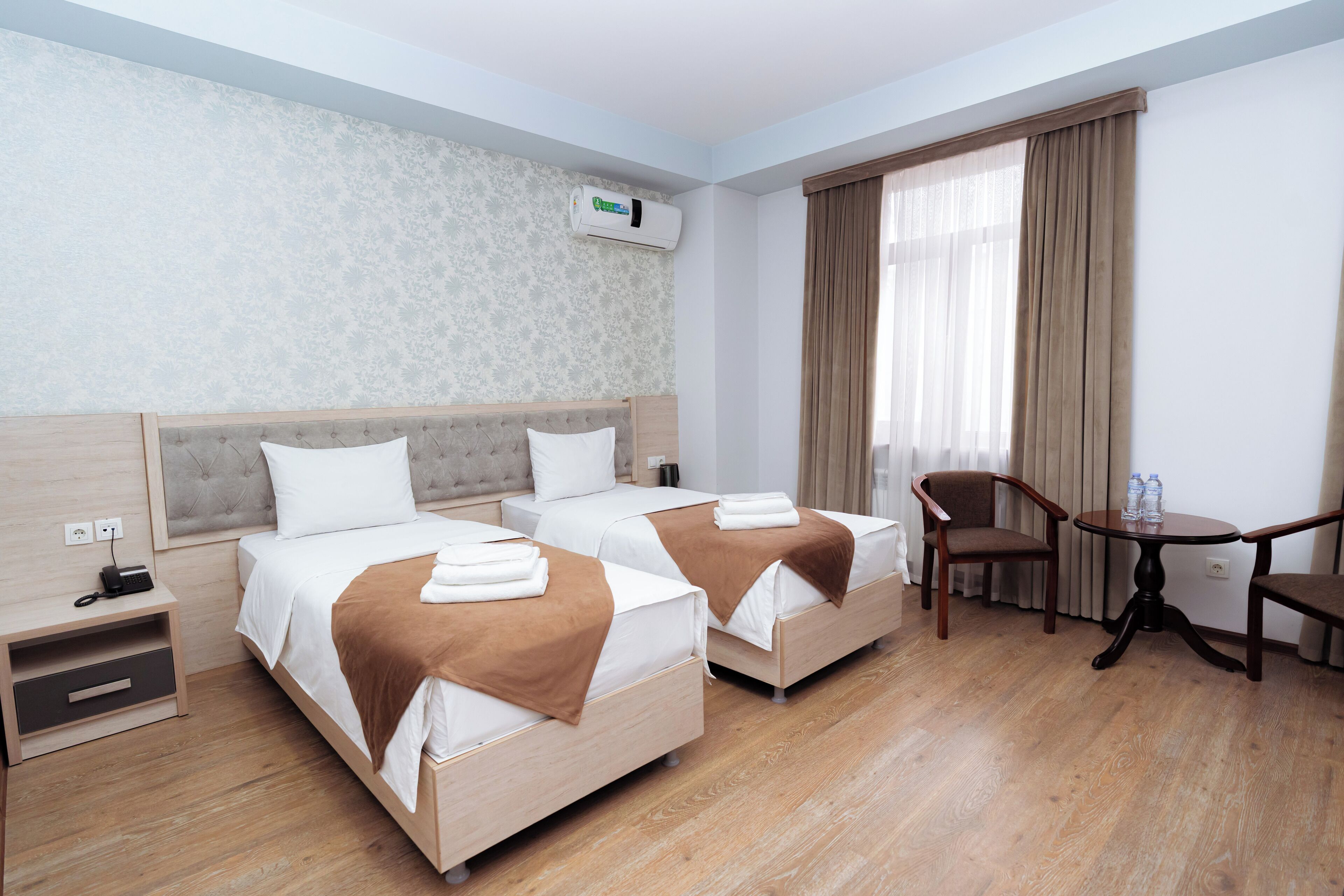 A hotel room with two beds, a small table, a chair, a nightstand, and an air conditioning unit.