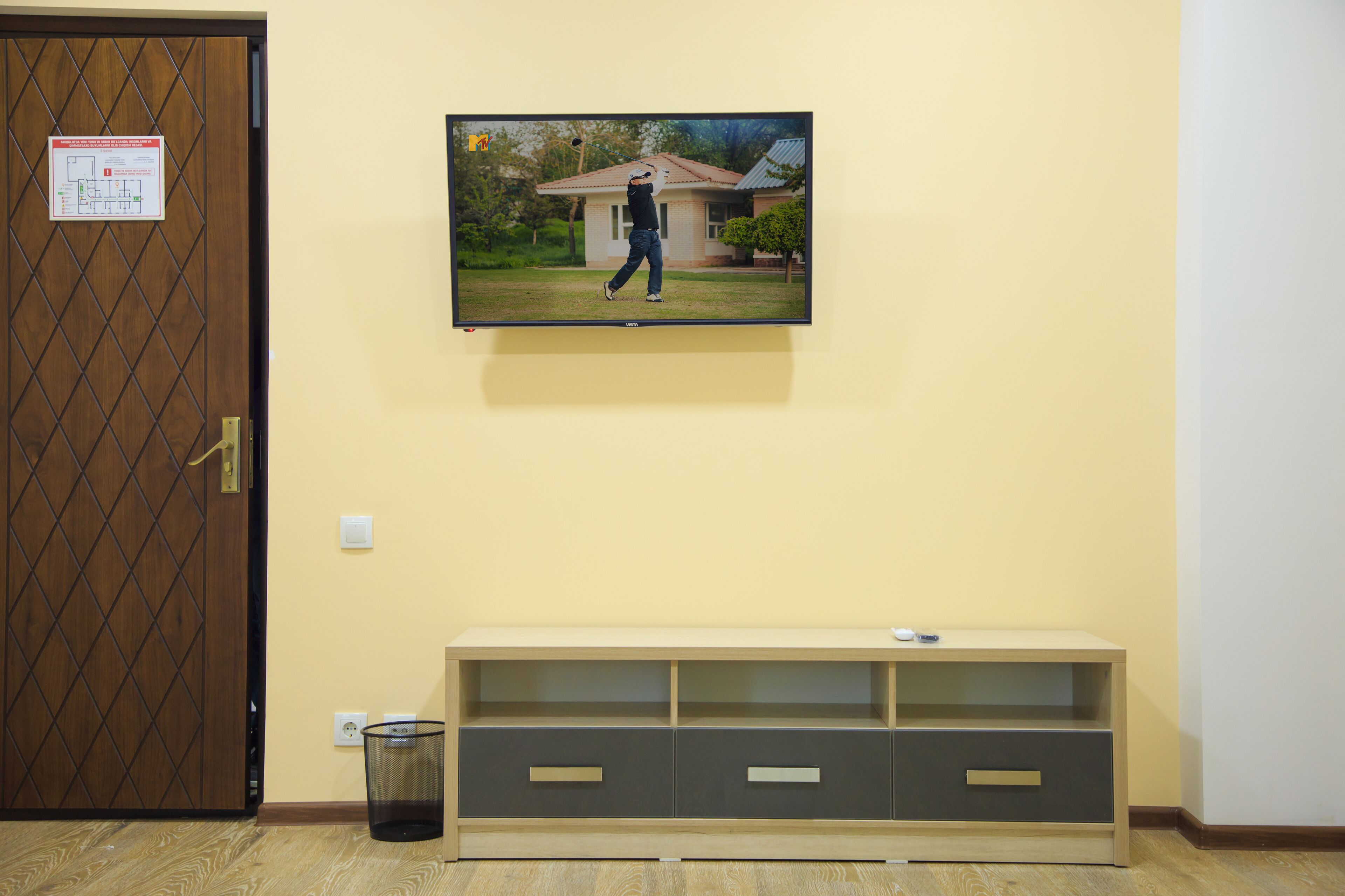 A room with a wooden door, a wall-mounted television displaying a golfer, a beige console table, and a trash bin.