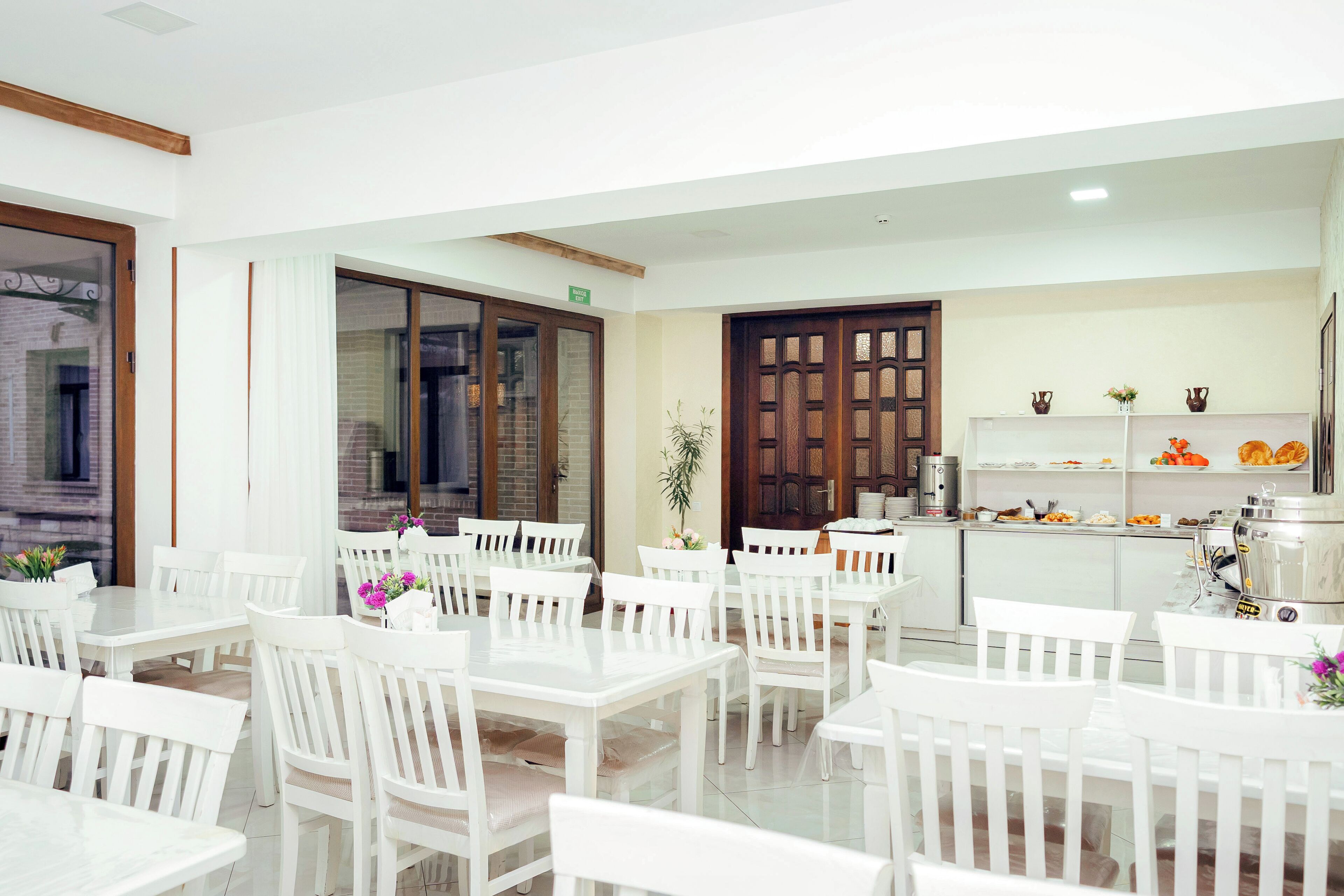 Un comedor con mesas y sillas blancas, un área de cocina con una cafetera y una puerta con un letrero verde.