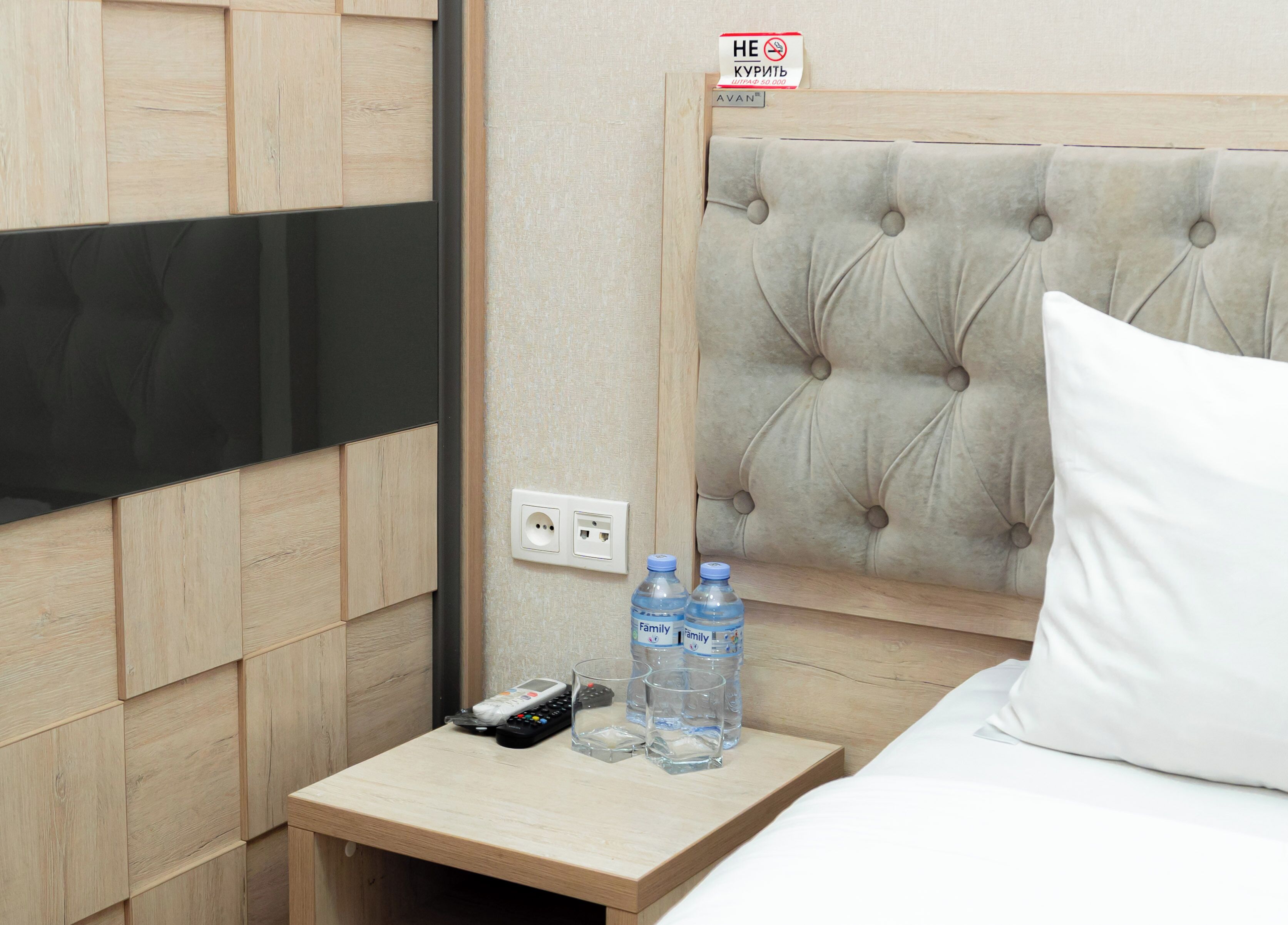 A hotel room with a wooden panel wall, a tufted headboard, a bedside table with a remote control, two water bottles, and a pillow.