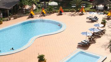 A swimming pool area with lounge chairs and umbrellas, surrounded by greenery and buildings in the background.