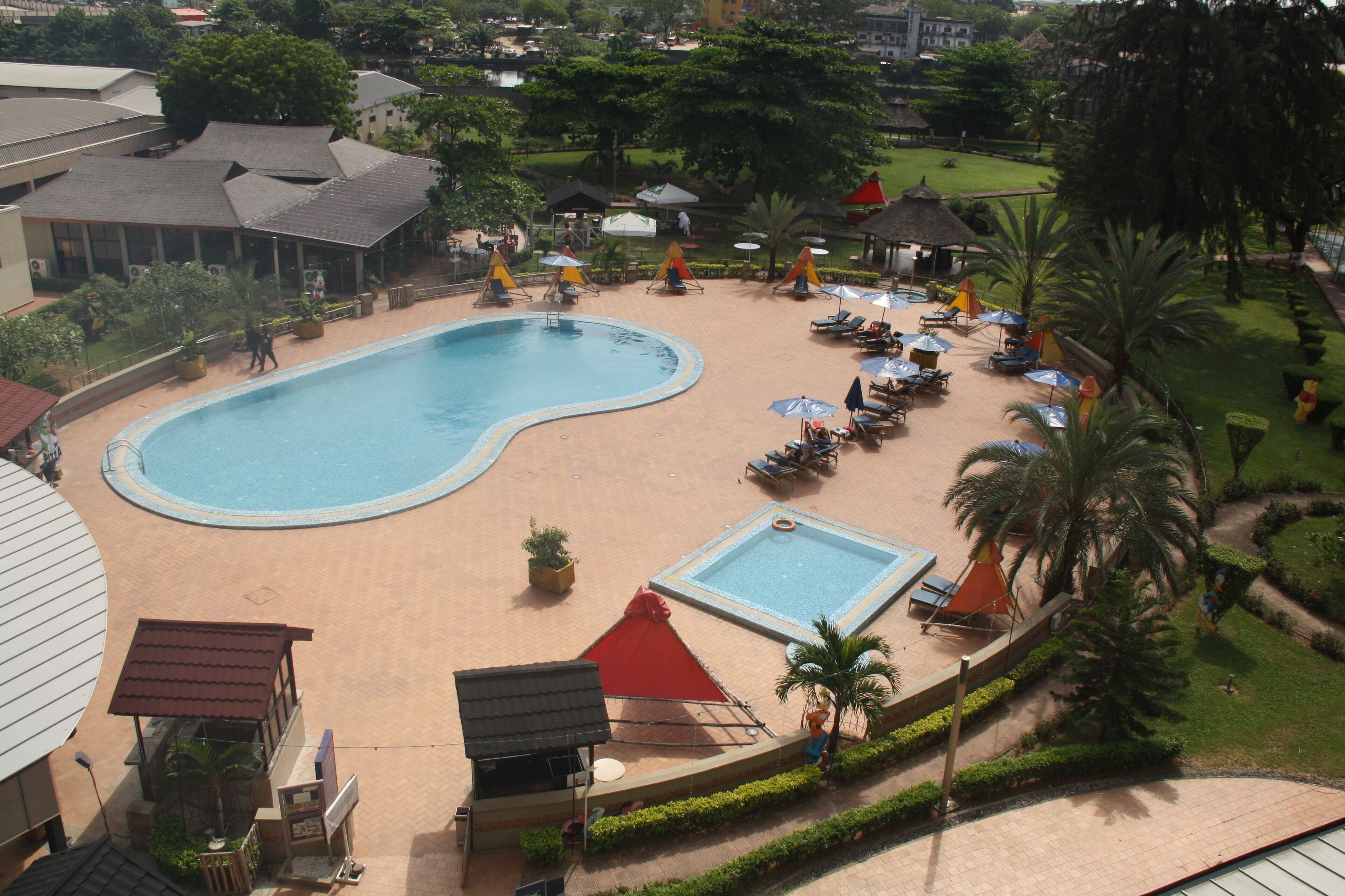 Zona de piscina con sillones de playa y sombrillas, rodeada de vegetación y edificios.