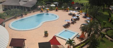 Zona de piscina con sillones de playa y sombrillas, rodeada de vegetación y edificios.