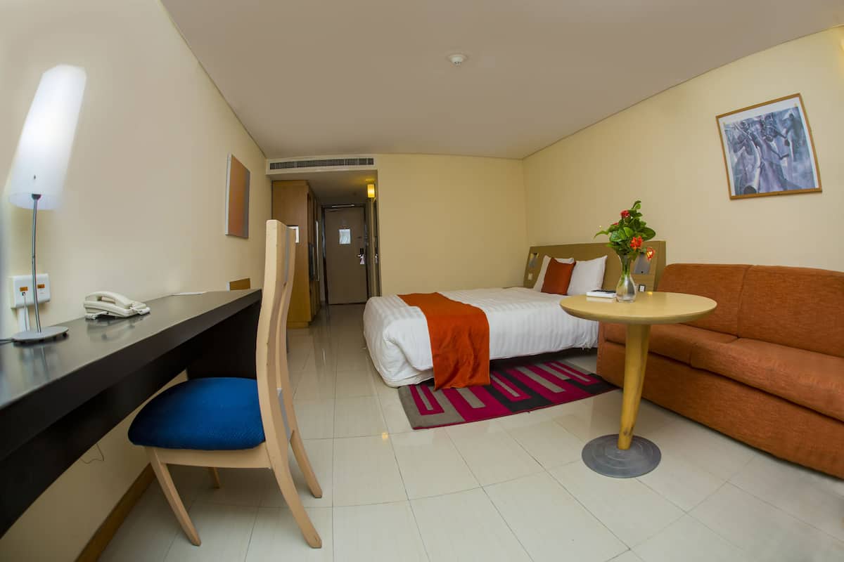 Ein Hotelzimmer mit einem Bett, einem Schreibtisch mit Telefon, einem Sessel, einer Couch, einem kleinen Tisch mit Vase und einem Bild an der Wand.