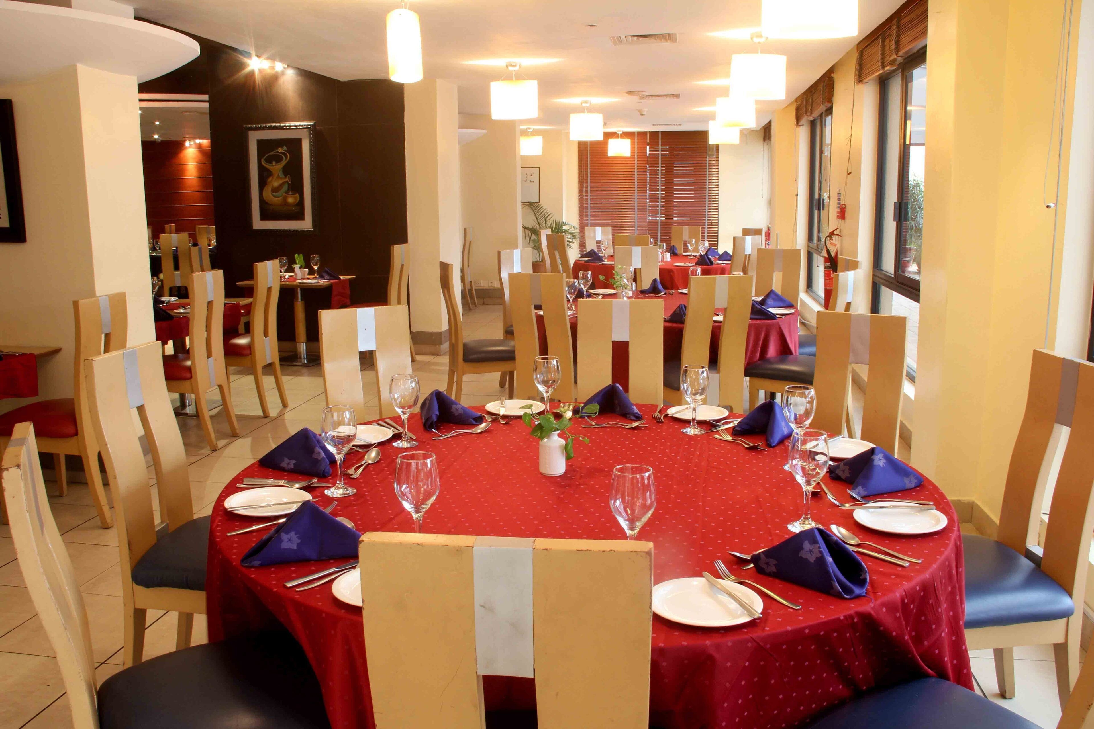 Un comedor con mesas redondas dispuestas para seis comensales, cada una con mantel rojo, platos blancos y servilletas azules.