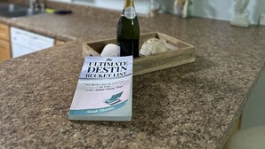 Un bancone della cucina con una bottiglia di champagne, un libro intitolato 'The Ultimate Destin Bucket List' e un vassoio di legno con conchiglie.