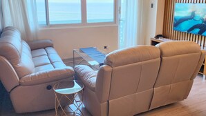 Un salon moderne avec un canapé beige, une petite table d’appoint et une grande fenêtre donnant sur la mer.