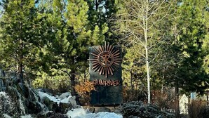 Et skilt med et sol-lignende design og teksten "SUNRIVER" i et naturområde med træer og en vej.