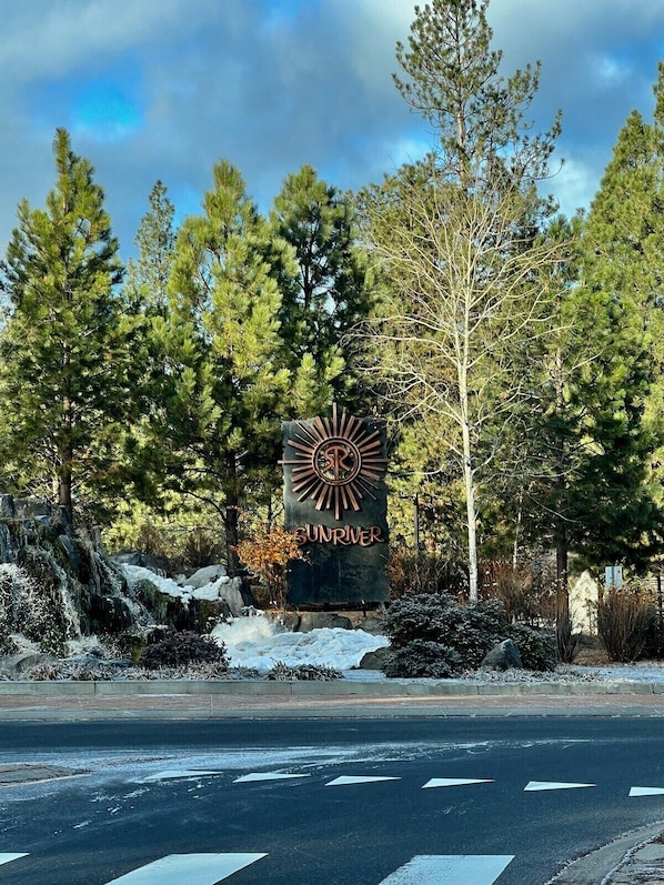 Um letreiro com um desenho semelhante ao sol e o texto 'SUNRIVER' em um ambiente natural com árvores e uma estrada.