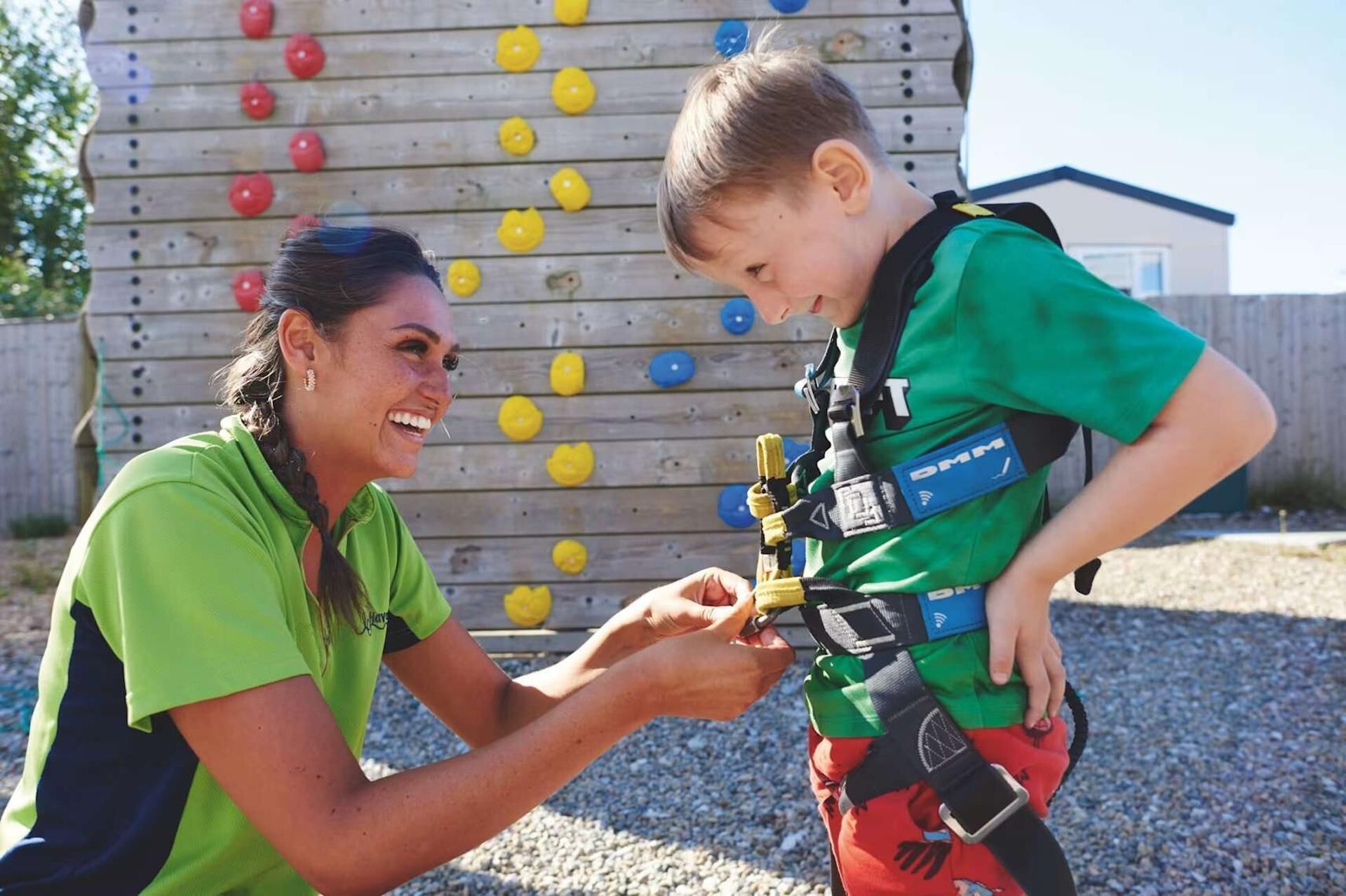 Une personne portant un t-shirt vert et un enfant portant un t-shirt vert avec un harnais, debout devant un mur d’escalade doté de prises rouges, jaunes et bleues.