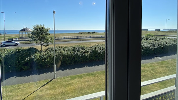 Vue depuis l’intérieur d’un bâtiment, donnant sur une route bordée d’une zone herbeuse et sur une étendue d’eau au loin.