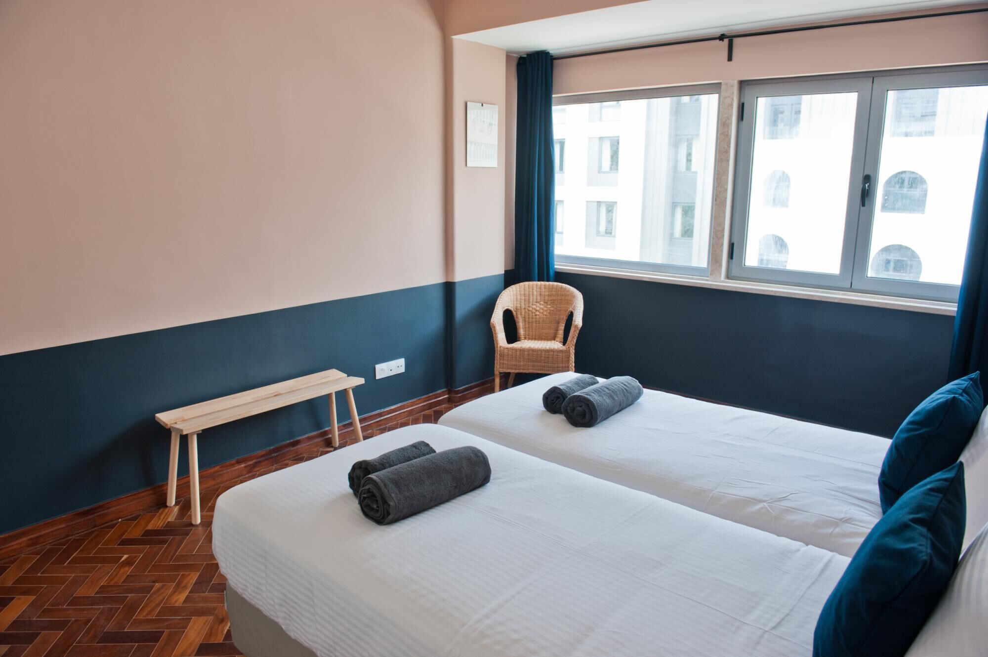 A hotel room with two beds, a bench, a wicker chair, and a window with a view of buildings.