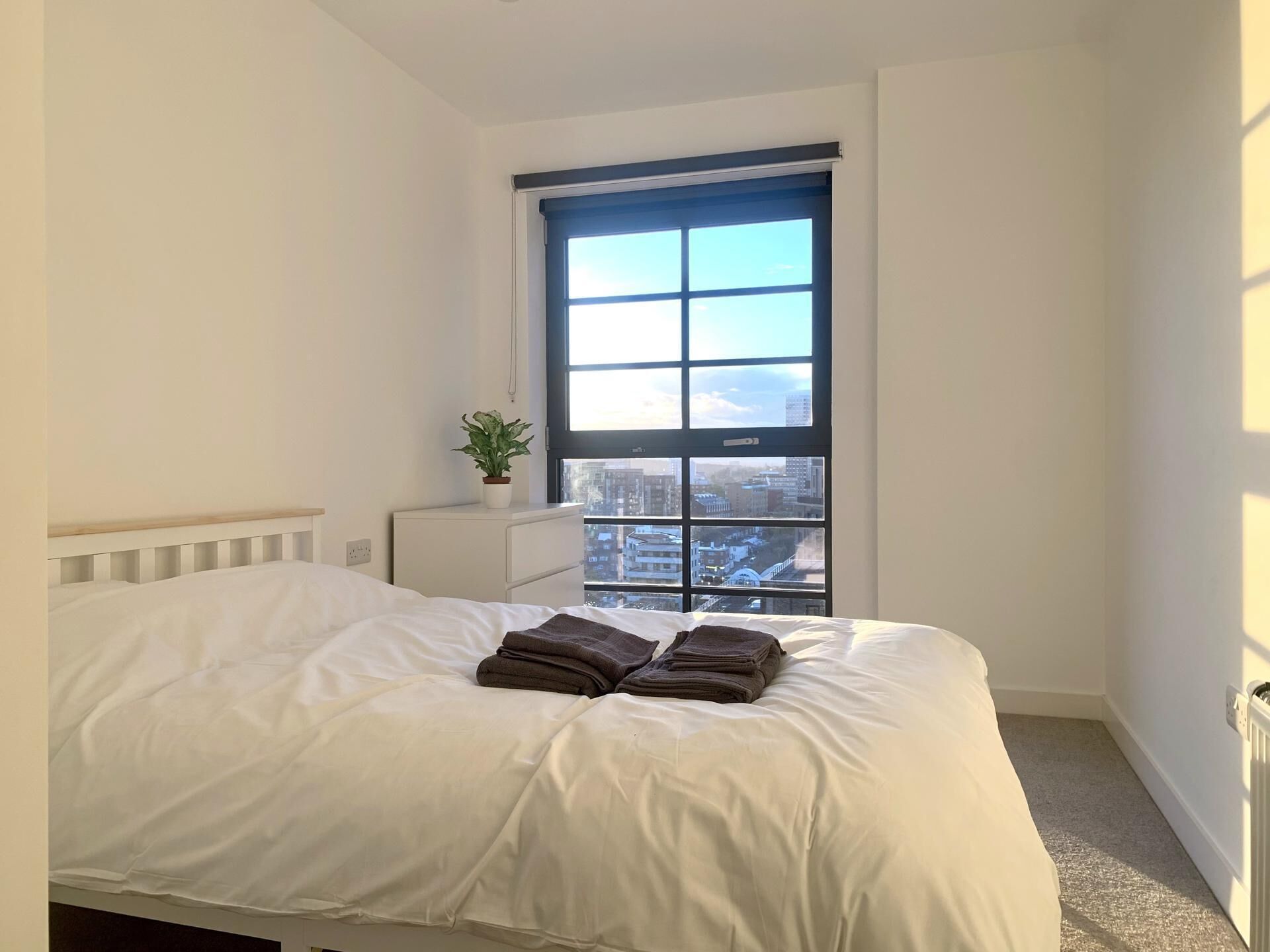 A bedroom with a large window, a bed with white bedding, a small bedside table with a plant, and a radiator on the wall.