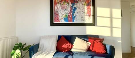 Una sala de estar con un sofá azul, cojines rojos y blancos, un cuadro abstracto enmarcado, una planta en maceta y una pequeña mesa de madera.