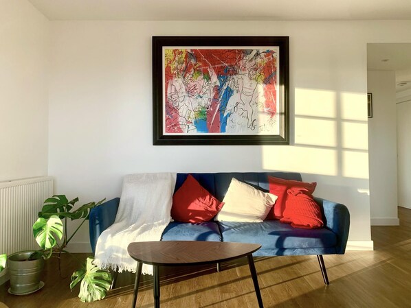 A living room with a blue sofa, red and white cushions, a framed abstract painting, a potted plant, and a small wooden table.