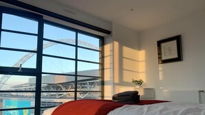 Quarto de hotel com uma grande janela, uma cama com roupa de cama branca e um cobertor vermelho, uma pequena mesa de cabeceira com uma planta e um quadro na parede.