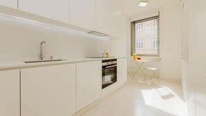 A modern kitchen with white cabinetry, a stainless steel sink, and an oven. There is a small table with a chair near a window. - Lady Madonna in Lisboa Ciudad (Lisbon)