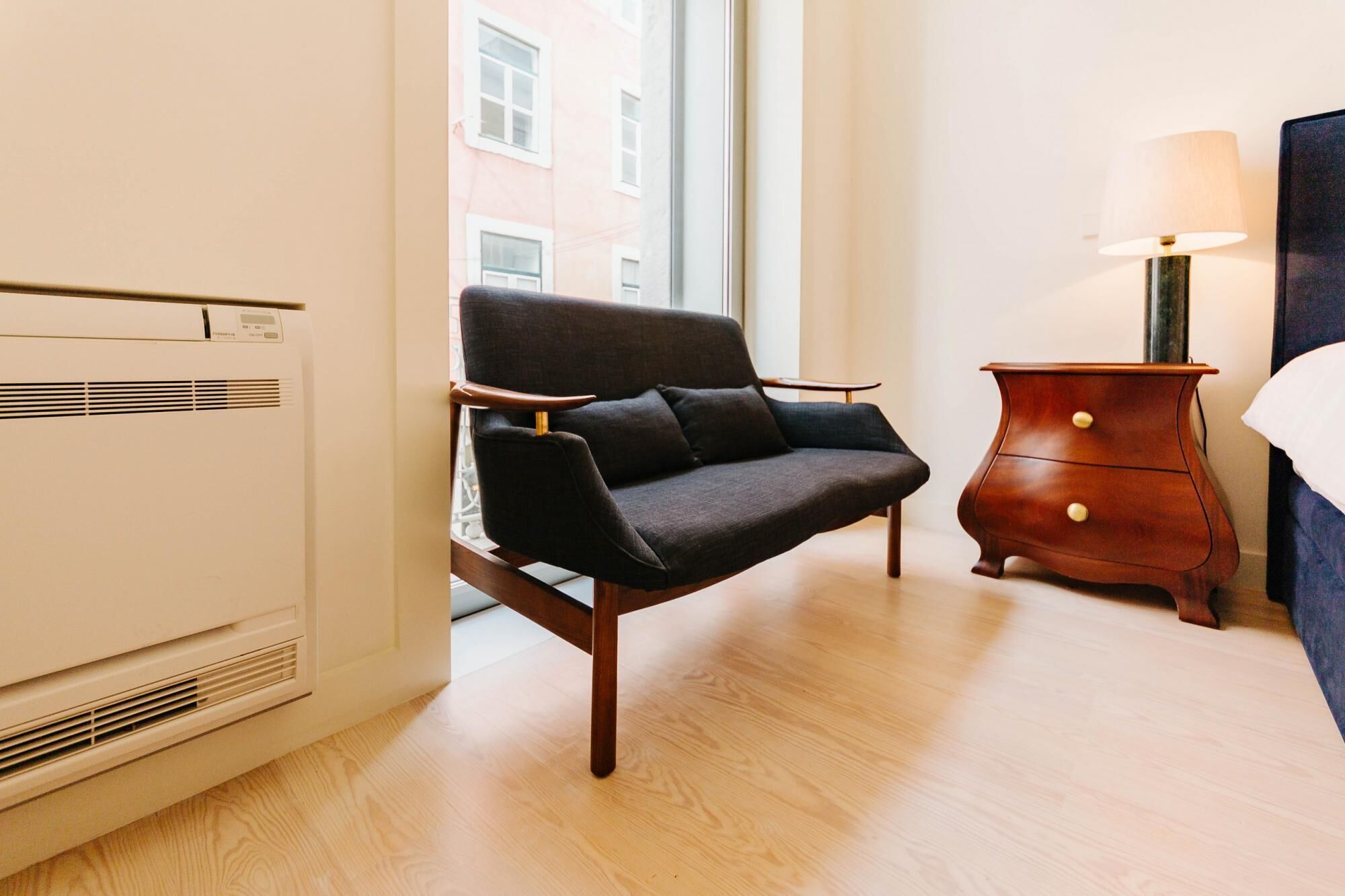 Une chambre d’hôtel moderne avec un canapé gris foncé, une table de chevet en bois avec une lampe et une grande fenêtre donnant sur les bâtiments environnants.