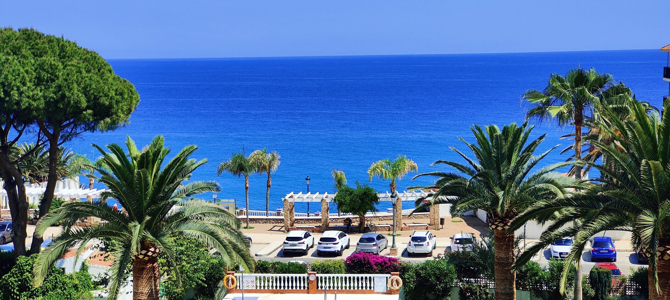 Vista costera con mar azul transparente, un estacionamiento con varios coches y palmeras.