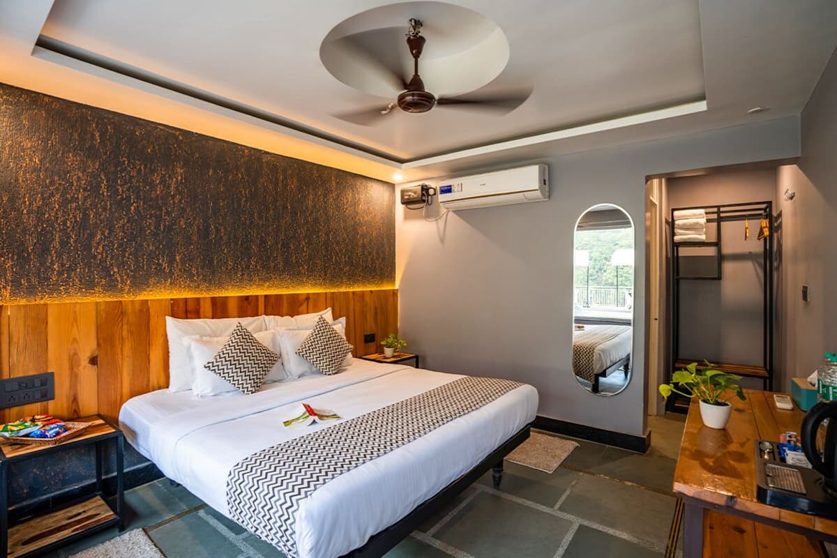 A modern hotel room with a wooden headboard, a bed with white bedding, a small bedside table with a lamp, a mirror, and a view of the outdoors.