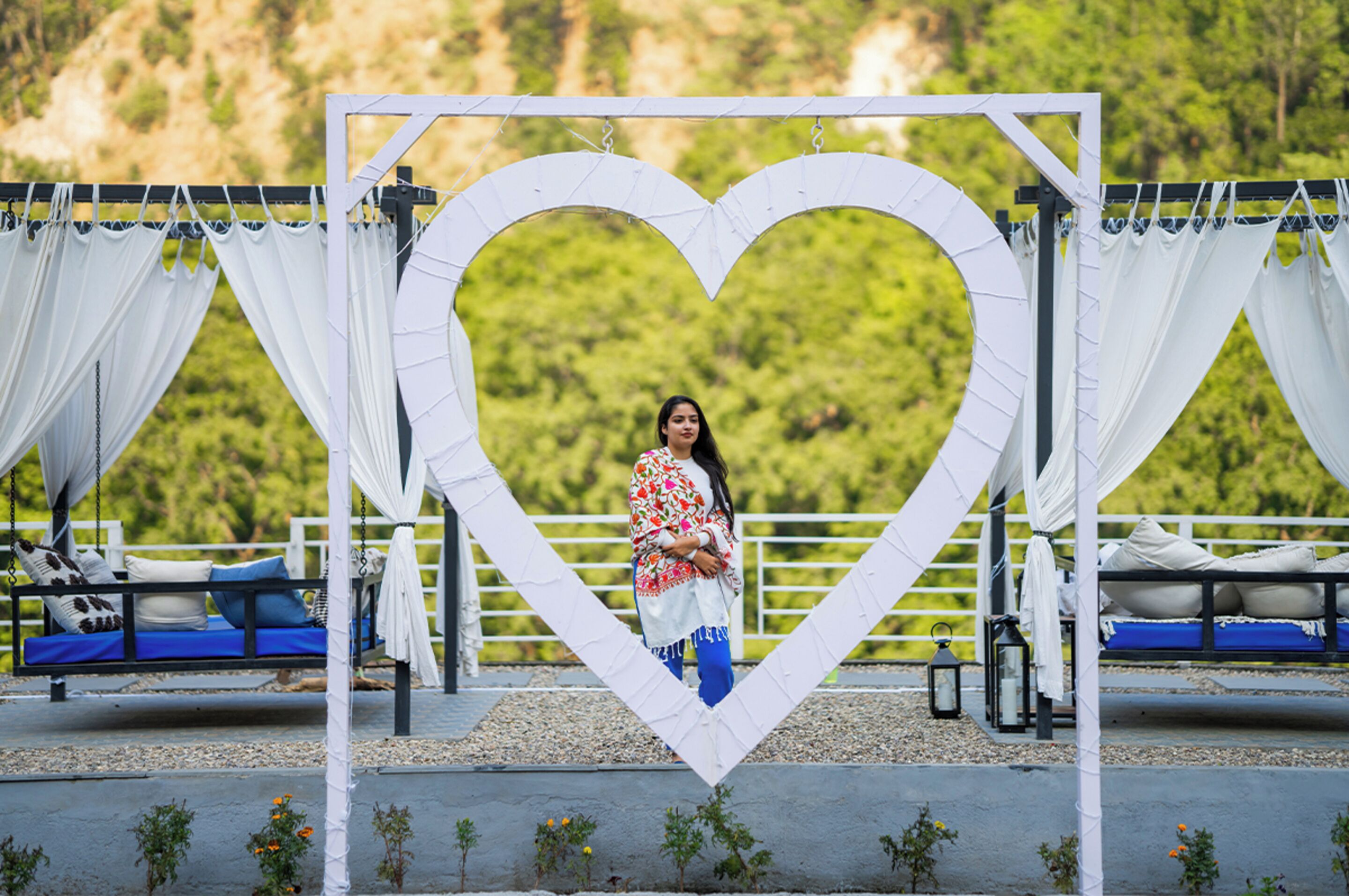 A person standing in front of a heart-shaped structure with white curtains, two beds with blue bedding, and a natural landscape in the background.