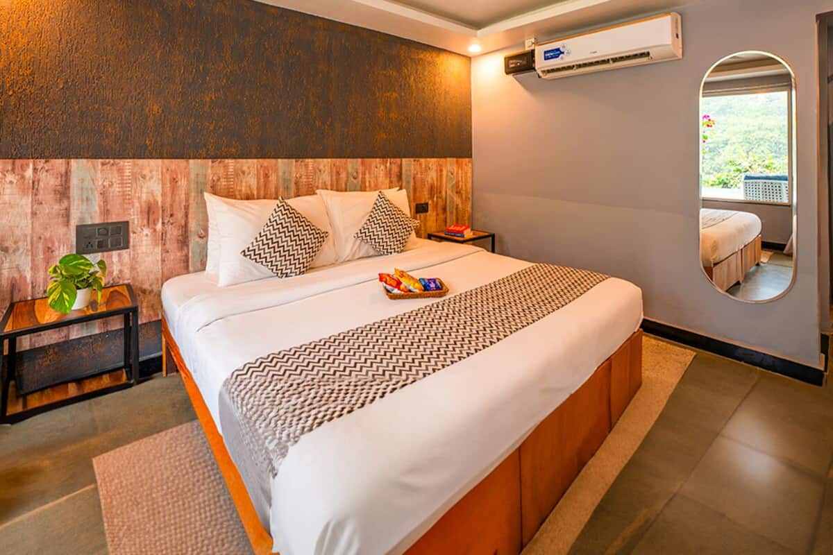 A modern bedroom with a wooden headboard, a large bed with patterned pillows, a bedside table with a plant, and a small round mirror.