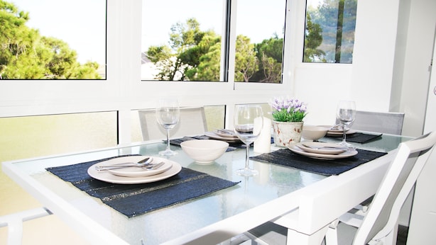 A dining table with a glass top, white chairs, and a view of trees through the windows. - Relax & Comfort in Lago Jardín • Garden Bungalow • Community Pool(35) (LOS BALCONES)
