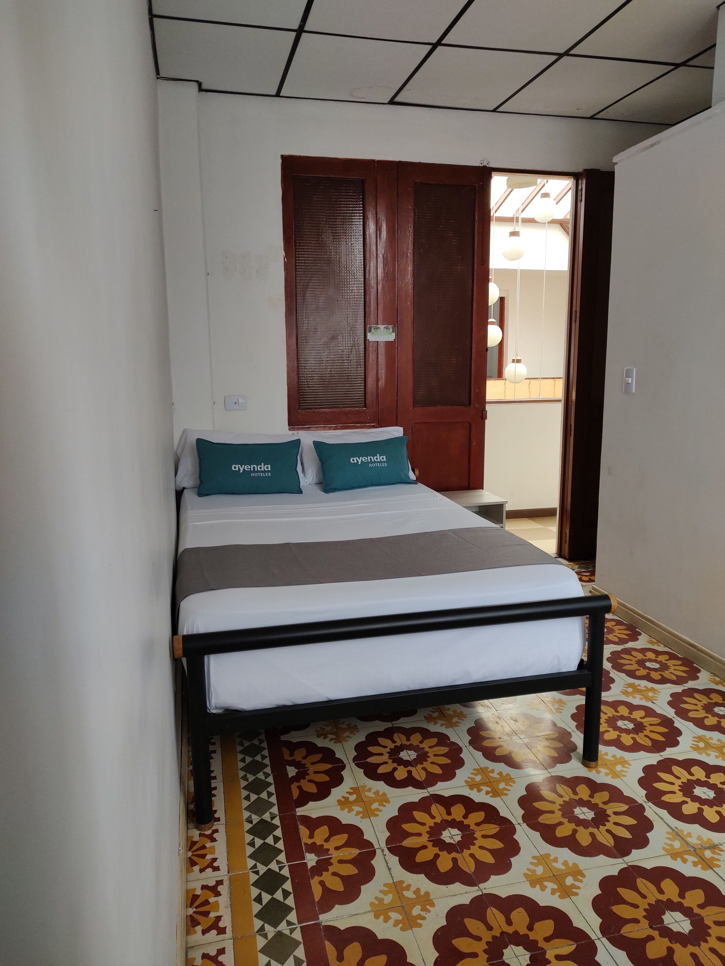A small, narrow hotel room with a bed, two pillows, a nightstand, and a door leading to a hallway.