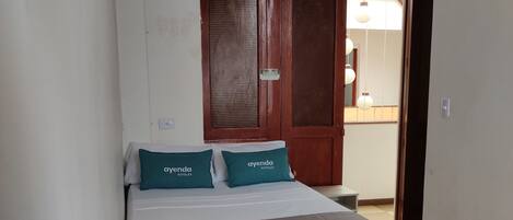 A small, narrow hotel room with a bed, two pillows, a nightstand, and a door leading to a hallway.