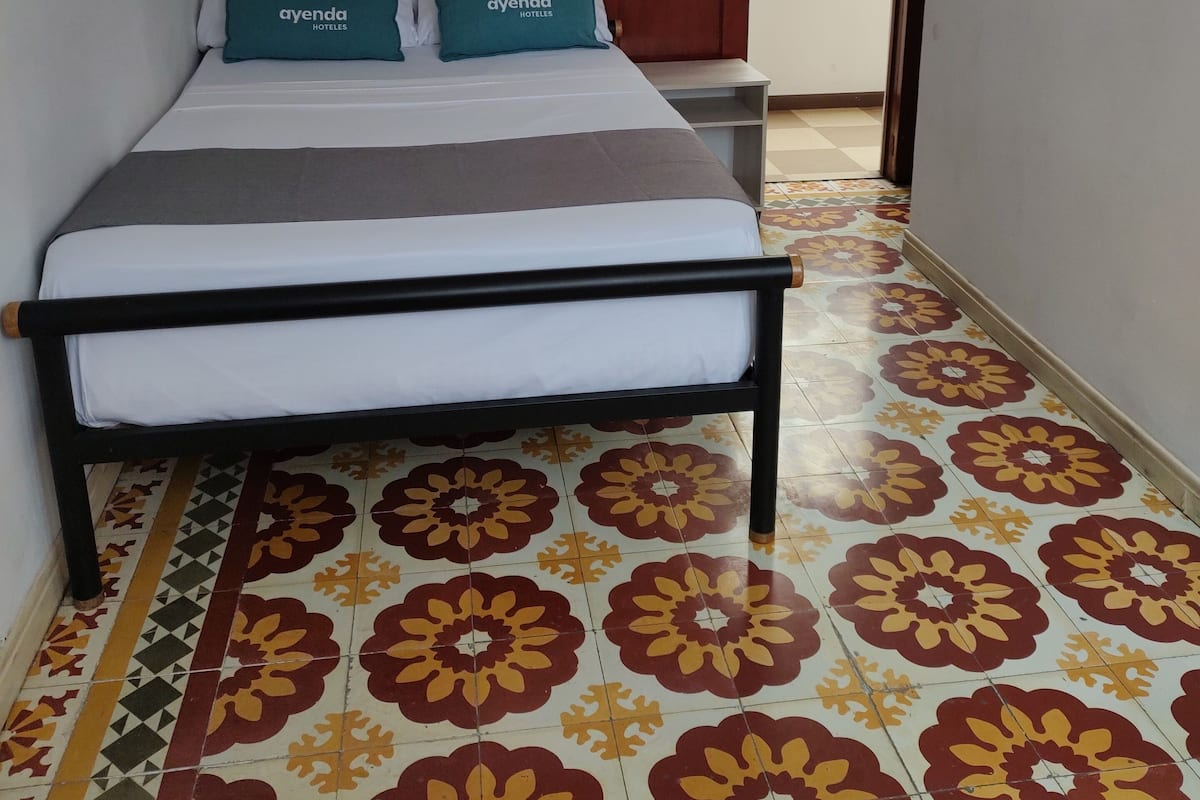 Ein Hotelzimmer mit einem Bett, zwei Kissen mit dem Schriftzug 'ayenda HOTELES', einer Holztür und einem gemusterten Boden.