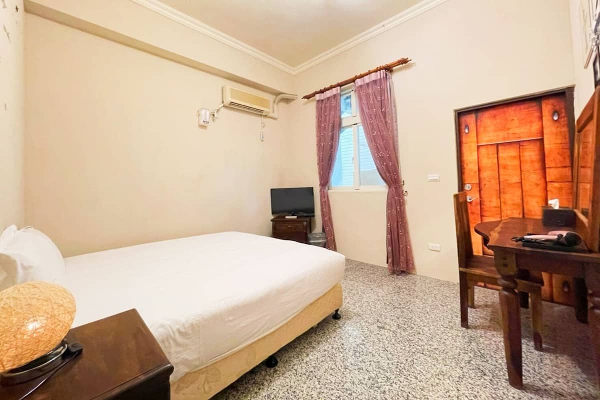 A hotel room with a bed, a desk, a chair, a television, and an air conditioning unit.