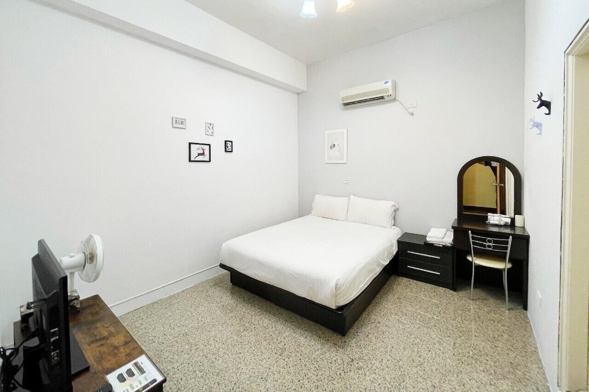 A bedroom with a bed, a desk, a chair, a mirror, and an air conditioning unit on the wall.