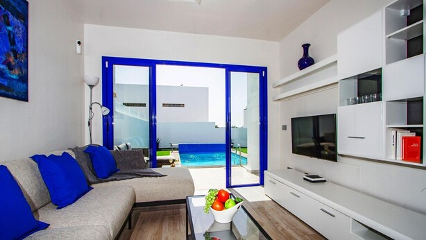 Un salon moderne avec un canapé, une table basse et un téléviseur. Une grande fenêtre offre une vue sur une piscine et un immeuble.