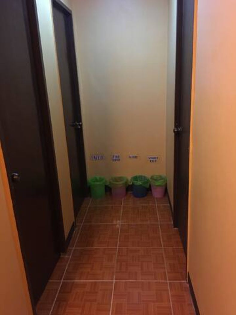 Een smalle gang met betegelde vloer en een deur aan de linkerkant. Tegen de muur staan vier vuilnisbakken op een rij.