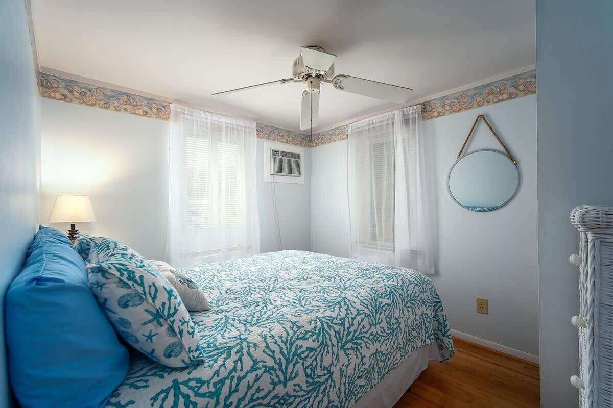 A bedroom with a bed, a ceiling fan, a window with curtains, a wall-mounted mirror, and a wicker chair.
