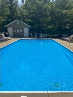 Une piscine rectangulaire dotée de deux plongeoirs, entourée de chaises longues et d’un petit bâtiment avec une porte et des fenêtres.
