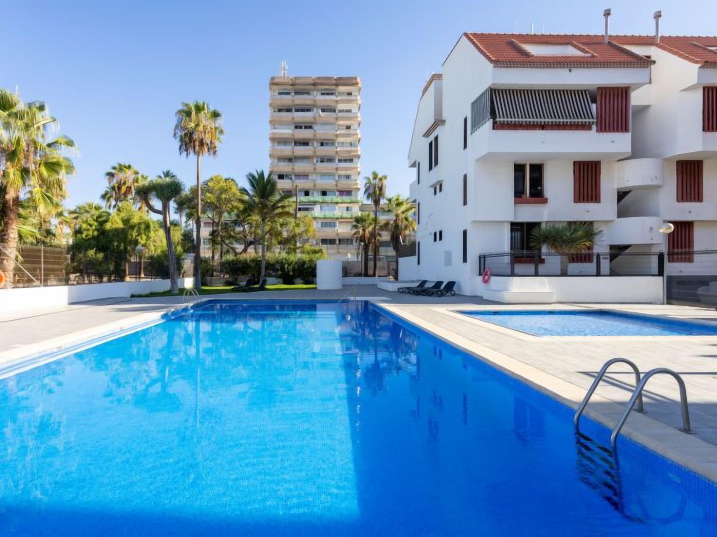 A swimming pool with a clear blue water surface, surrounded by a white building with balconies and red shutters.