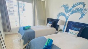 Habitación de hotel con dos camas, cada una con una colcha azul y blanca, un cojín con la palabra 'BEACH' y un mural de una palmera.