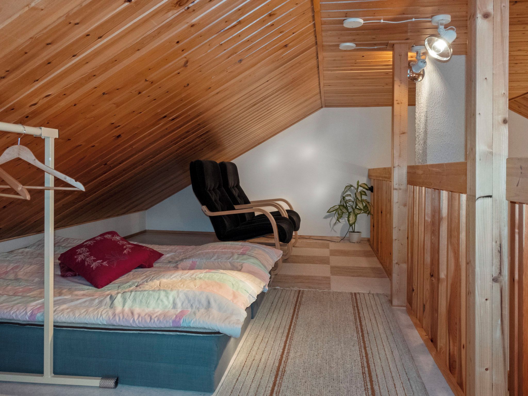 A bedroom with a sloped ceiling, a bed with a colorful quilt, a black chair, a small plant, and a wooden bench.