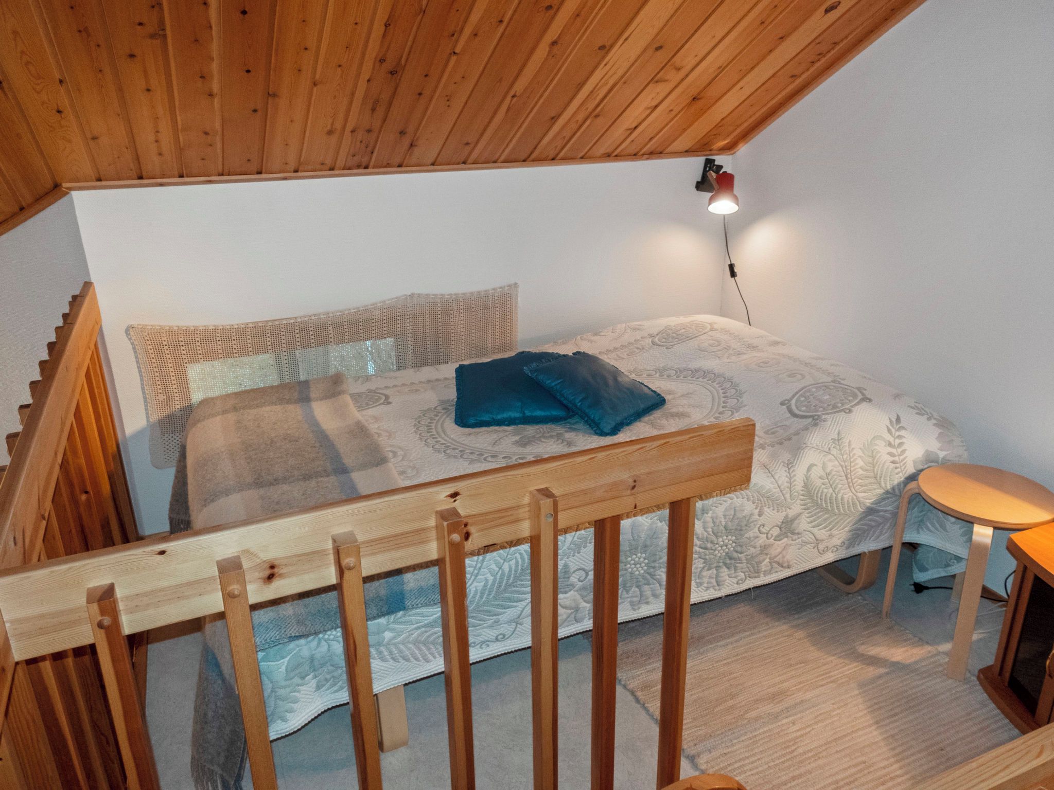 Un dormitorio con techo de madera, una cama con ropa de cama estampada, un taburete pequeño y una lámpara de pared.