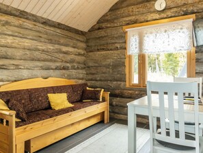 Interior de una cabaña de madera con un sofá de madera, una silla blanca y una ventana con cortina estampada.