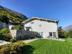 Une maison moderne à deux étages, dotée d’un mur de soutènement en pierre, d’une pelouse bien entretenue et surplombant un paysage montagneux.