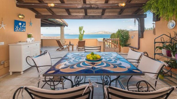 A covered outdoor dining area with a view of the sea, a table with a decorative tablecloth, and several chairs. - Valentina by Interhome (Cannigione)
