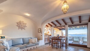 A spacious living area with a sofa, dining table, and chairs, a decorative ceiling light, and a view of the sea through large windows. - Valentina by Interhome (Cannigione)