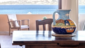 A wooden table with a colorful ceramic pitcher in front of a window overlooking a body of water and a distant hill. - Valentina by Interhome (Cannigione)