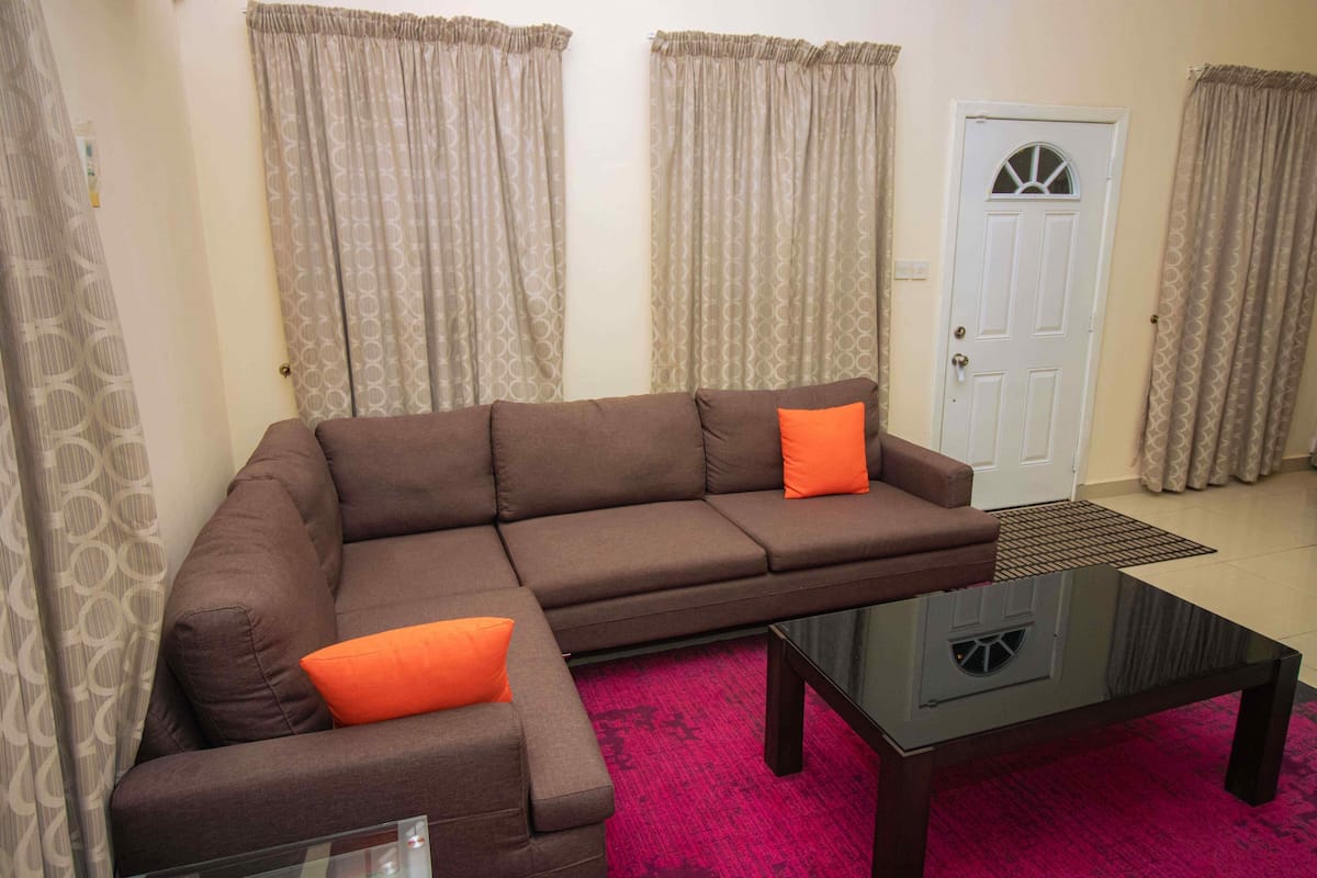 A living room with a brown sectional sofa, orange cushions, a glass coffee table, patterned curtains, and a white door with a window.