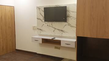 Un baño con puerta de madera, pared de mármol, televisor montado y un estante pequeño con un control remoto.