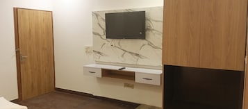 A bathroom with a wooden door, a marble wall, a mounted TV, and a small shelf with a remote.
