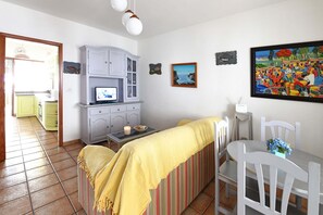 A cozy living room with a sofa, a dining table, and chairs. There's a television on a cabinet, a painting on the wall, and a window with a view of the outside.
