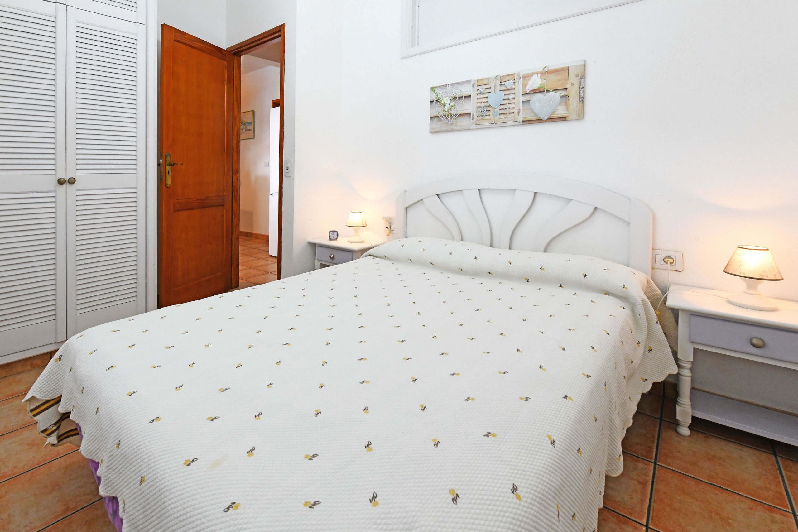 A bedroom with a bed, two bedside tables, a nightstand, a wooden door, and a wall-mounted decoration.